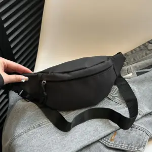 Schwarze Bauchtasche aus Nylon liegt auf einer Jeansjacke, Hand hält den Gurt