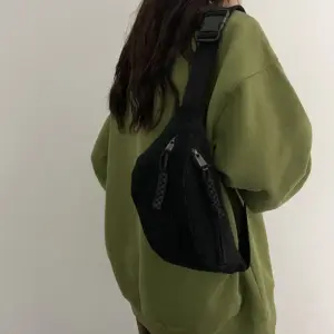 Schwarze Cord-Bauchtasche mit Reißverschlüssen, auf dem Rücken über grünem Pullover getragen