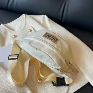 Cremefarbene Bauchtasche mit Etikett und breitem Gurt auf einer hellen Jacke liegend