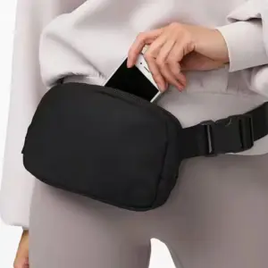 Schwarze Gürteltasche mit Reißverschluss, in der Hand mit Handy, Person in hellem Oberteil